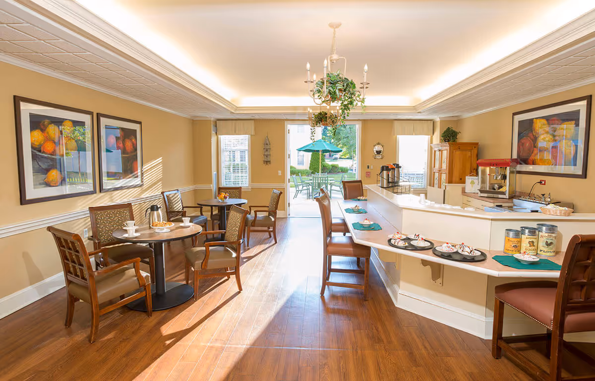 A bright and inviting dining area with wooden floors, beige walls, and a white ceiling with recessed lighting. There are several round tables with chairs, each set with cups and plates. A counter with bar stools displays plates of desserts and coffee containers. A chandelier with hanging plants is centered above the counter. Large windows and a door at the back open to an outdoor patio with tables and a green umbrella.
