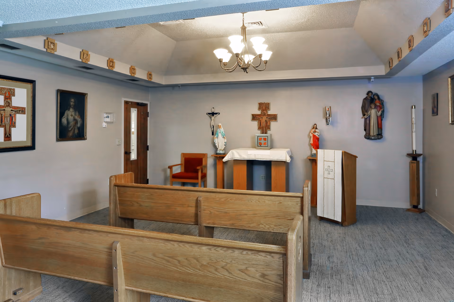 Small chapel room with wooden pews facing an altar. The altar has religious statues and crosses on the wall behind it. A chandelier hangs from the ceiling, and religious paintings are on the walls.