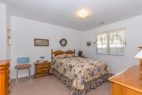 A bedroom with a wooden bed frame and a floral bedspread in shades of beige and purple. There is a wooden nightstand with a lamp and decorative items next to the bed. A wooden dresser is visible on the right side of the image, and a small blue chair is placed near the nightstand. The room has white walls with framed pictures and a decorative wreath above the bed. A window with lace curtains allows natural light into the room.