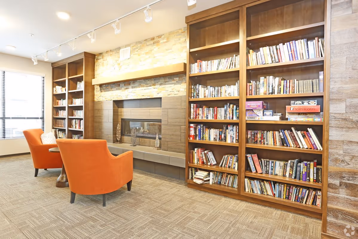 A cozy reading area with two orange armchairs facing a modern fireplace set in a stone and tile wall. Flanking the fireplace are tall wooden bookshelves filled with books and board games. A large window with blinds allows natural light into the room, which has carpeted flooring and track lighting on the ceiling.