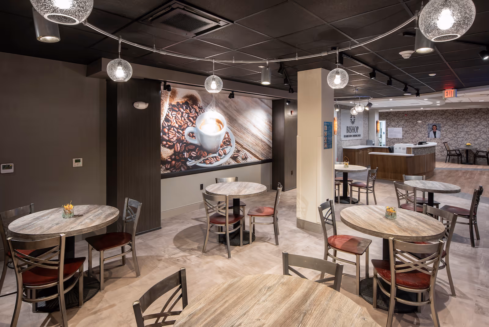 Modern communal dining area with round wooden tables and chairs, pendant lights, and a large coffee-themed mural on the wall.