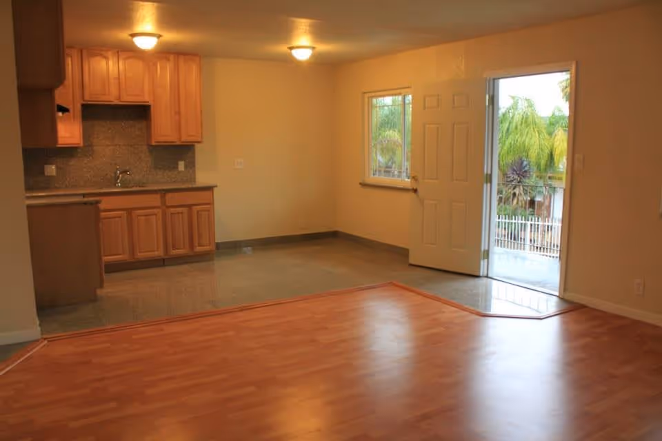 Interior view of a living space with a small kitchen area featuring wooden cabinets and a countertop on the left side. The room has a combination of wood and tile flooring, ceiling lights, a window, and an open door leading to an outdoor area with visible greenery.