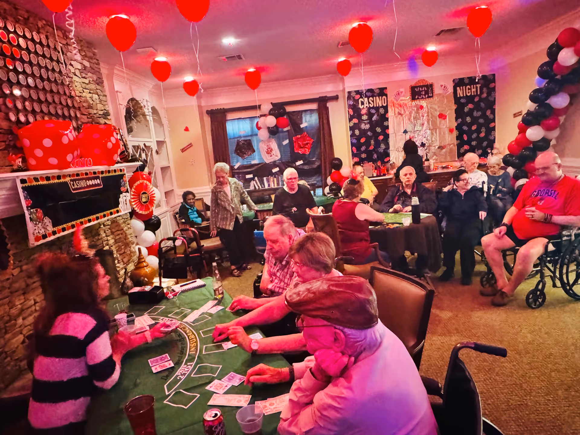 A group of elderly people gathered in a decorated room playing card games at a green felt table. The room is festively adorned with red, black, and white balloons, large red dice decorations, and banners that read 'CASINO NIGHT'. Some people are seated in wheelchairs while others sit or stand around the table and in the background.