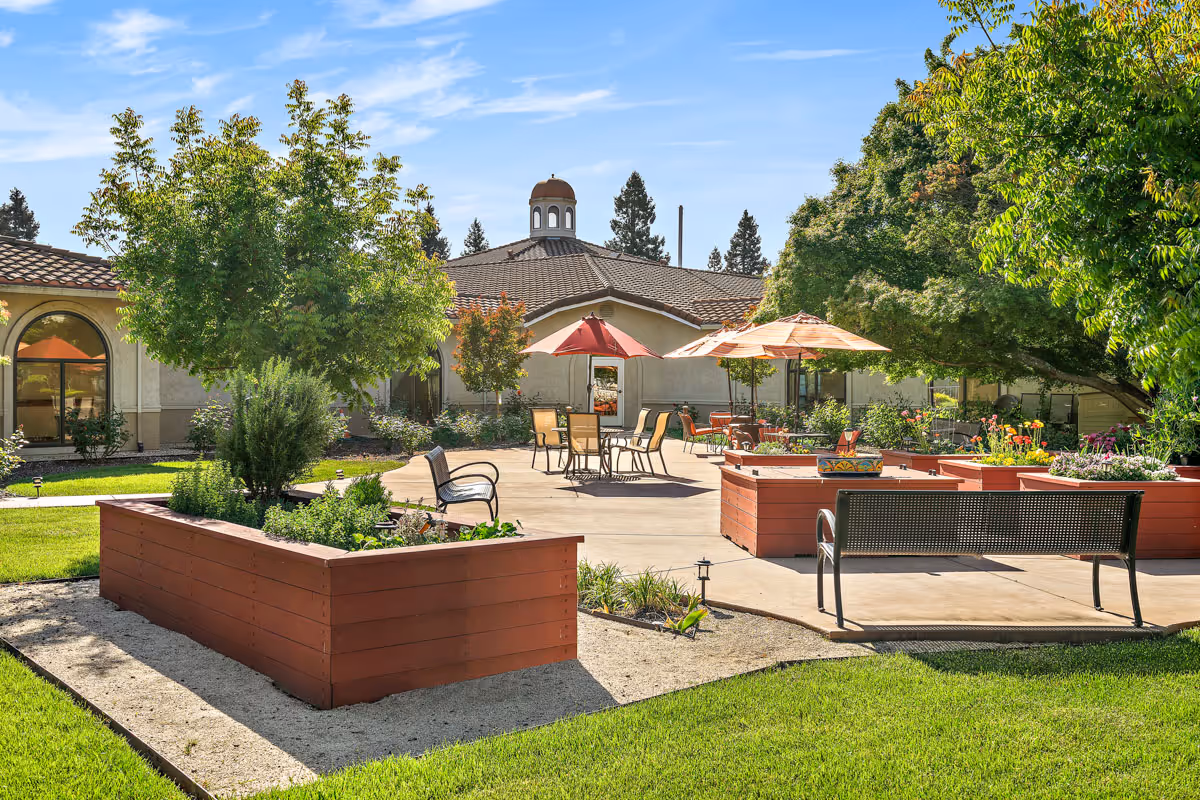 Outdoor courtyard area with raised garden beds, patio tables with umbrellas, chairs, benches, and surrounding greenery including trees and shrubs under a clear blue sky.