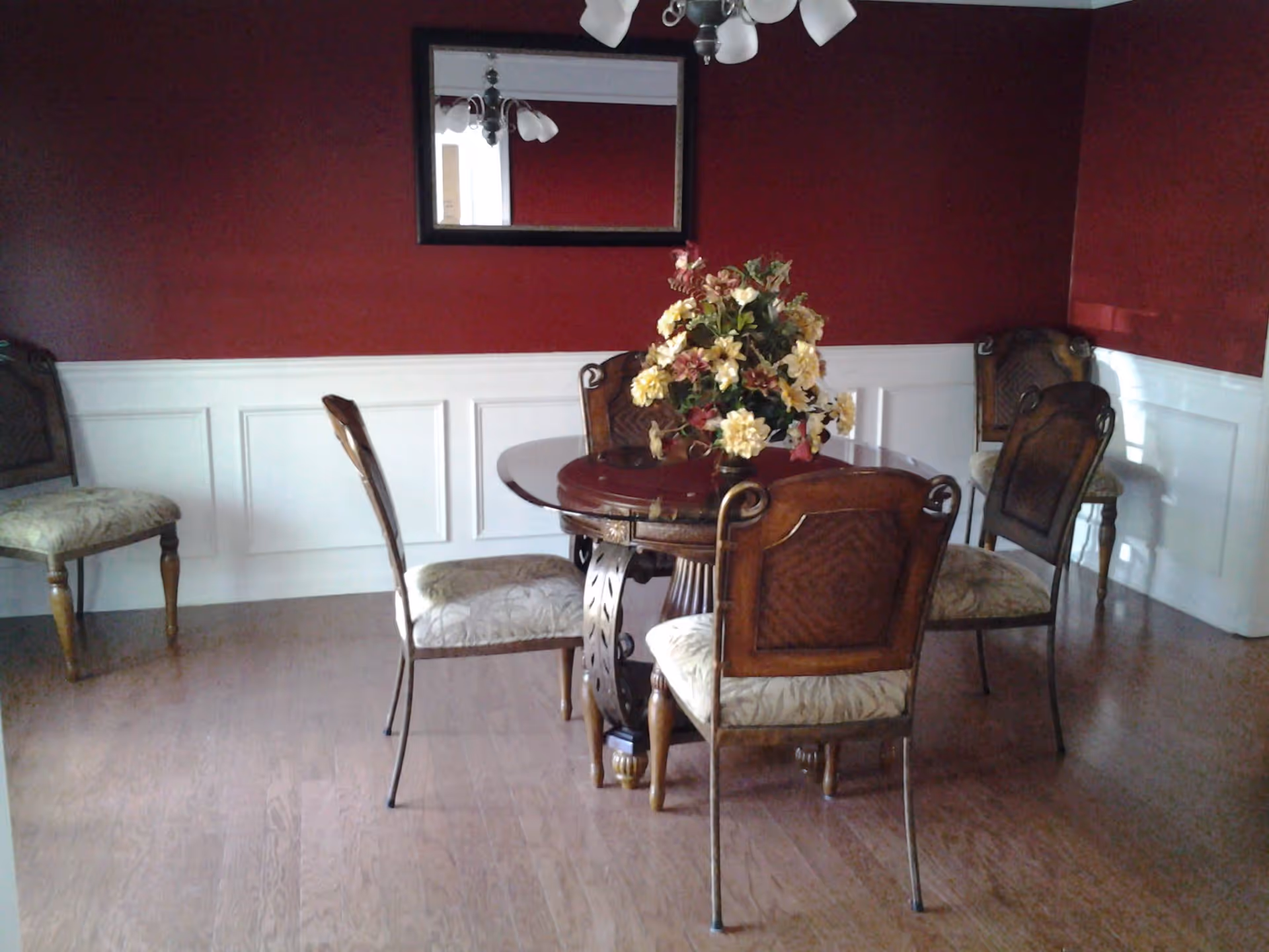 Dining room with a round wooden table topped by a floral centerpiece and several upholstered chairs against red upper walls and white wainscoting.