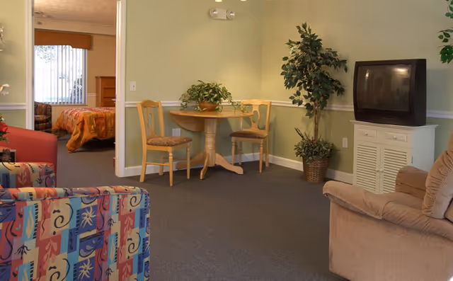A cozy living area with a small round wooden table and two chairs, a TV on a white cabinet, a potted plant, and a patterned armchair. In the background, a bedroom with a bed covered in an orange and red bedspread is visible through an open doorway.