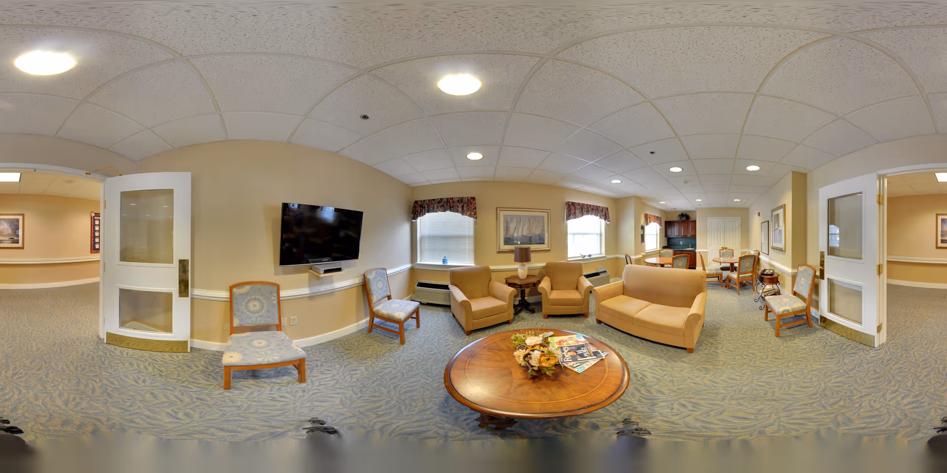 A spacious senior living common area with beige walls and carpeted floor. The room features a round wooden coffee table with magazines and a decorative centerpiece, surrounded by beige upholstered armchairs and a sofa. Two patterned chairs are placed near a wall-mounted flat-screen TV. The room has two windows with floral valances, framed artwork on the walls, and a small dining area with a table and chairs in the background. Double glass doors open to a hallway.