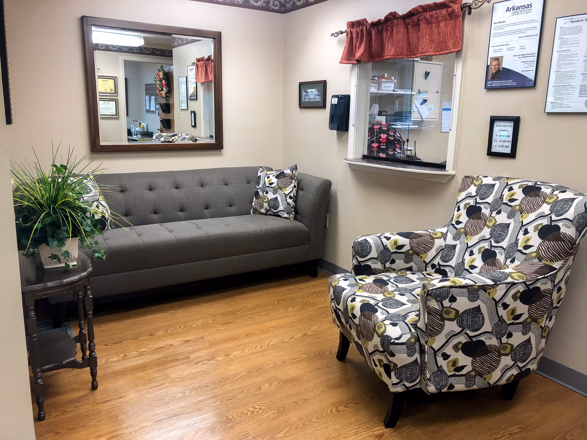 Reception waiting area with a gray tufted sofa, patterned armchair, side table with a plant, and a reception window with a mirror on the wall.