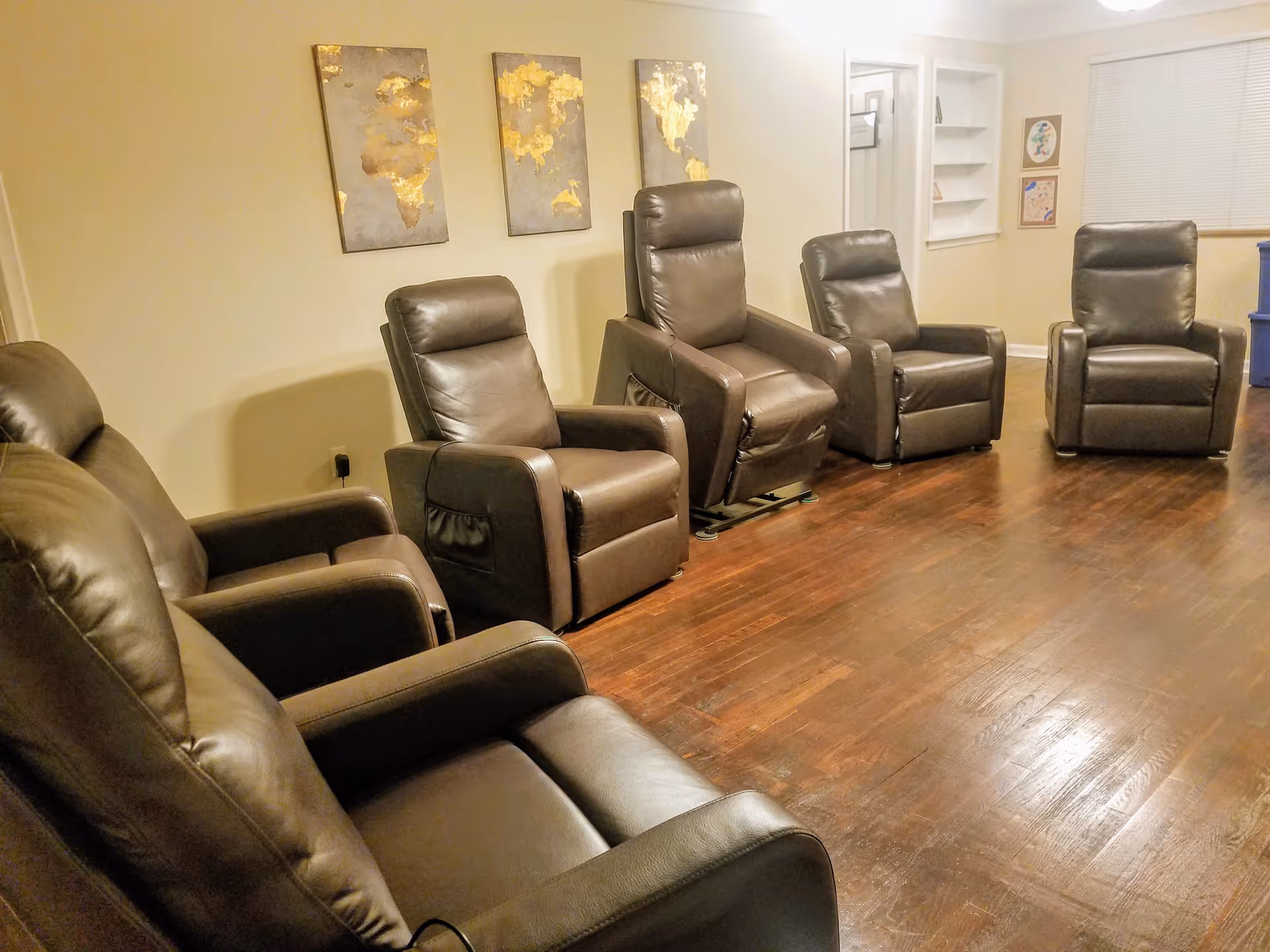 Row of brown leather recliner chairs arranged in a communal living room with hardwood floors and wall art.