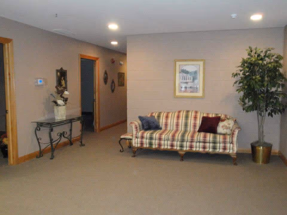 Interior view of a living area with a plaid patterned sofa adorned with three pillows, a framed picture hanging on the wall above the sofa, a potted artificial tree to the right, and a decorative glass table with a flower arrangement on the left. The walls are painted beige with wooden trim around the doorways and baseboards.
