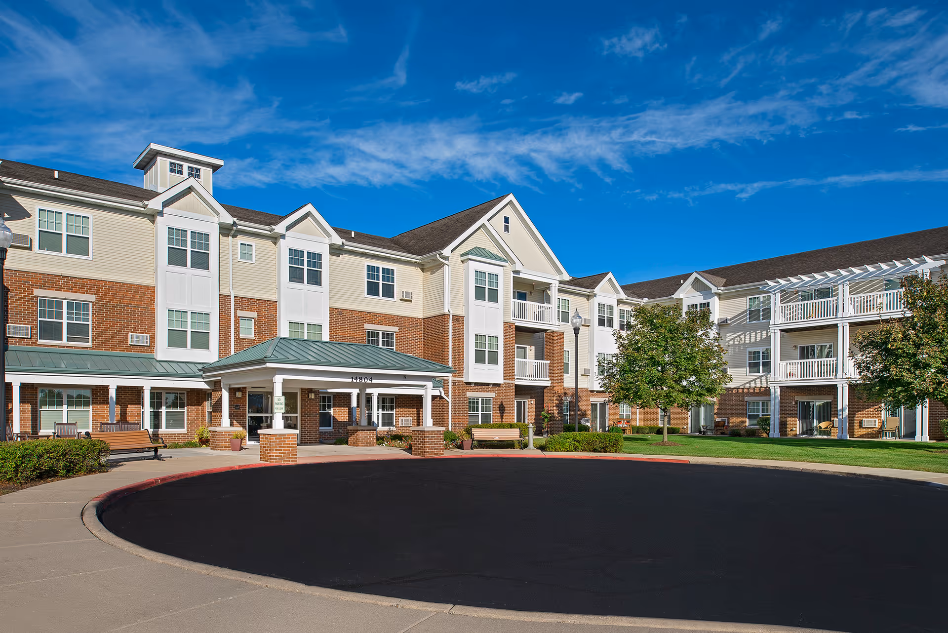Exterior view of a three-story senior living facility building with a covered entrance, multiple windows, balconies, and well-maintained landscaping under a blue sky with some clouds.