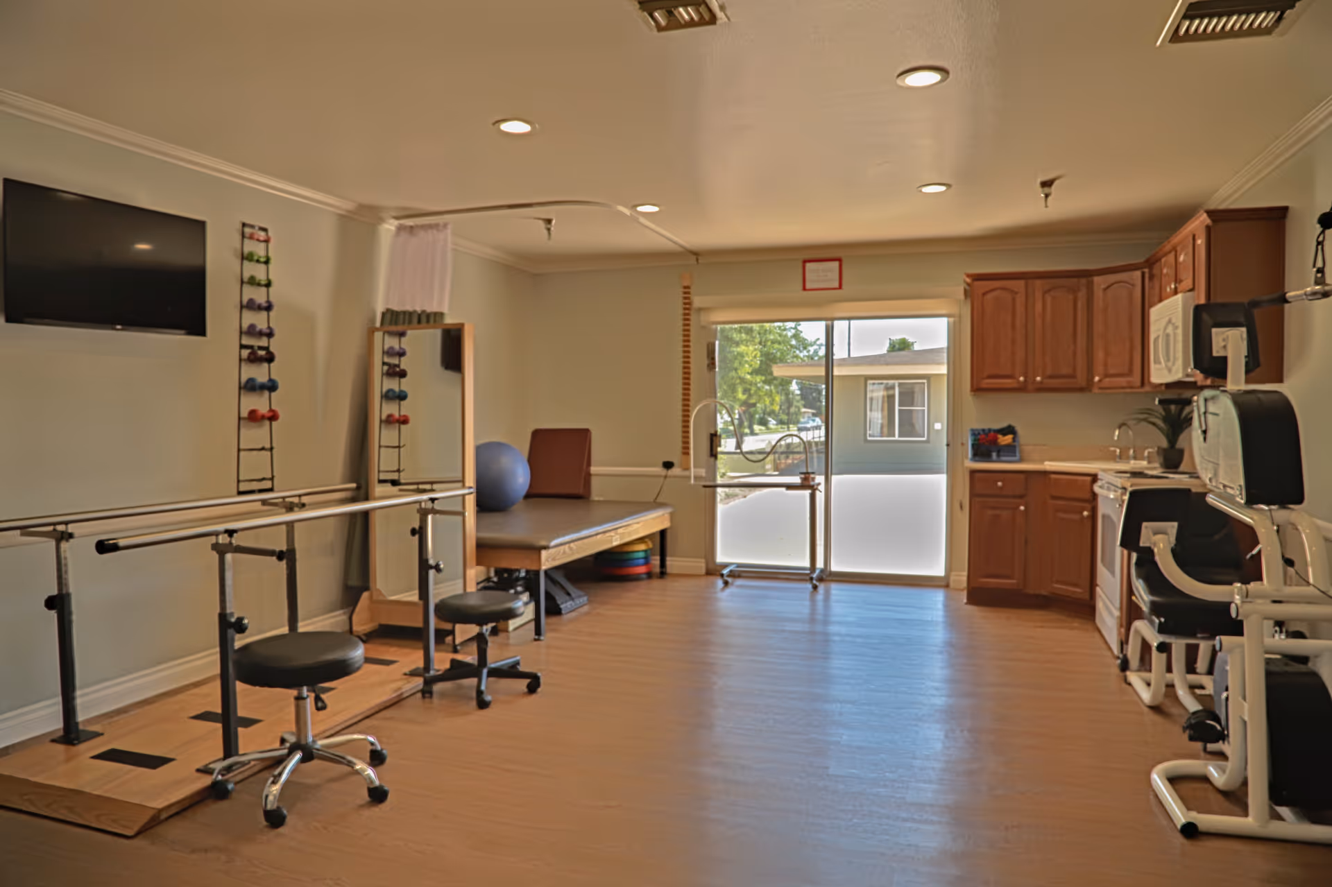 A rehabilitation or physical therapy room with parallel bars for walking exercises, two stools, a therapy table with a blue exercise ball, a rack of colorful hand weights, a large mirror, and exercise equipment. There is a kitchen area with wooden cabinets, a microwave, and a sink on the right side. A large window and glass door provide natural light and a view of the outside.