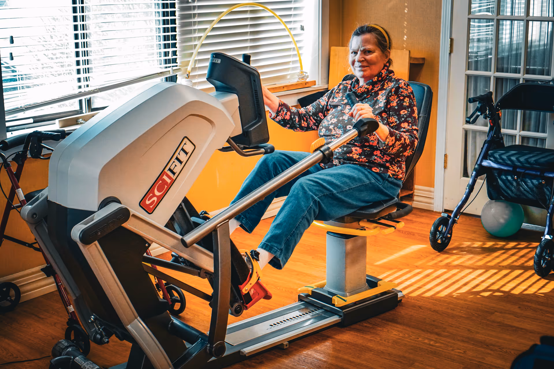 An elderly woman using a Schift exercise machine in a well-lit room with wooden flooring. She is seated and holding the machine's handles while pedaling with her feet. Behind her, there are windows with blinds, a walker, and a door with glass panels.