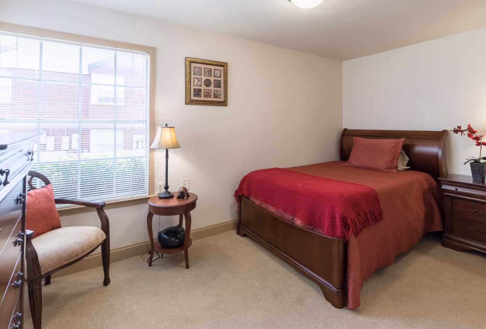 A cozy bedroom with a wooden bed covered in a red bedspread and matching pillow. Next to the bed is a wooden nightstand with a red orchid plant. A small round wooden side table with a lamp and a radio sits beside a window with blinds. There is also a wooden armchair with a red cushion and a wooden dresser. A framed picture hangs on the wall above the side table.