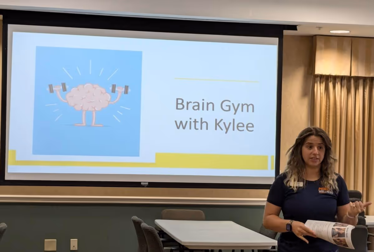 A woman stands in front of a projection screen in a meeting room. The screen displays a slide with a cartoon brain lifting dumbbells and the text 'Brain Gym with Kylee'. The room has tables and chairs, and beige curtains cover the windows.
