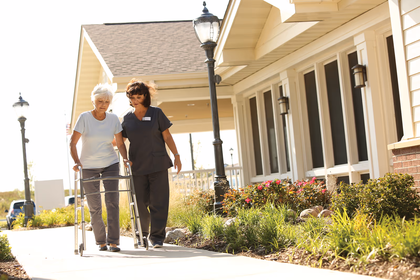 An elderly woman using a walker is being assisted by a caregiver as they walk along a sidewalk outside a building with beige siding and large windows. There are street lamps and well-maintained landscaping with green plants and flowers along the path.