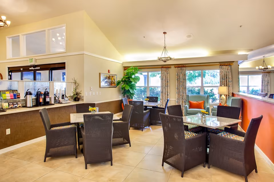 A bright and welcoming common area in a senior living facility with several tables and wicker chairs arranged for seating. There is a counter with coffee dispensers and a rack of coffee supplies on the left side. Large windows with patterned curtains let in natural light, and there are decorative plants and framed artwork on the walls. The space has tiled floors and warm, neutral-colored walls with an orange accent wall on the right.