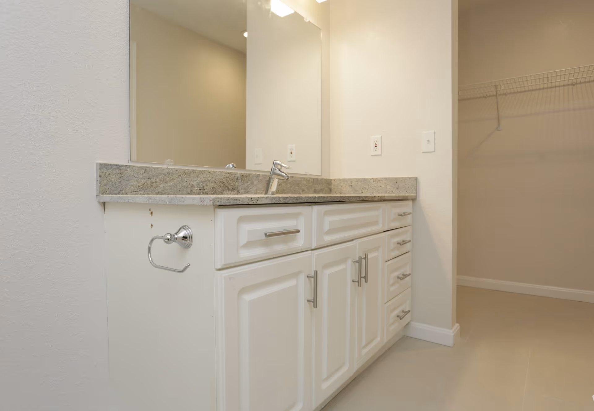 A bathroom vanity with a granite countertop, a single faucet, and white cabinets with silver handles. A large mirror is mounted above the vanity. To the right, there is an open walk-in closet with a wire shelf. The walls and floor are light-colored.