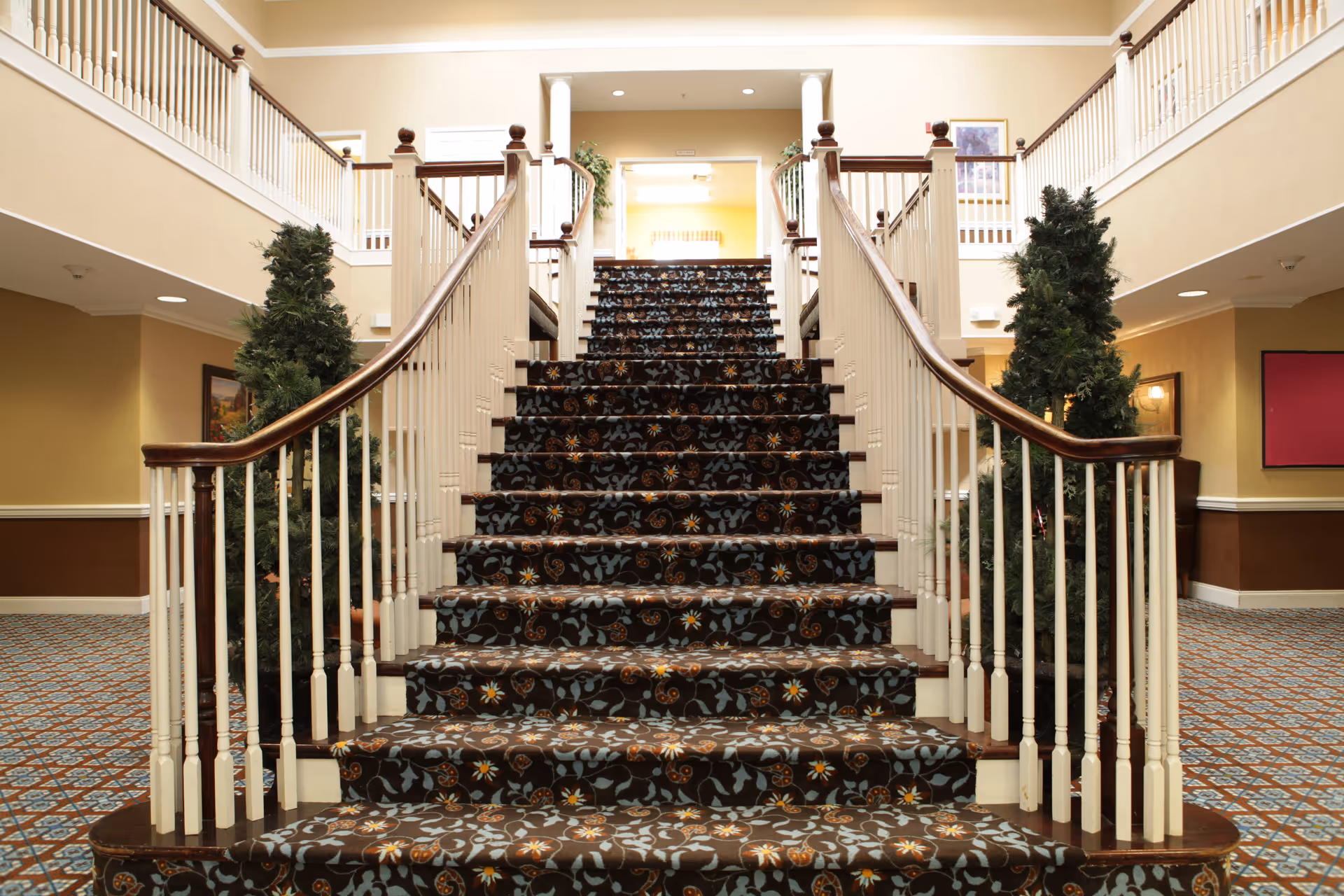 Carpeted staircase with floral pattern leading up to a second floor in an interior space. The staircase has white railings with dark wooden handrails. There are two potted evergreen plants on either side of the staircase at the bottom. The walls are painted beige with white trim, and there is a patterned carpet on the floor surrounding the staircase. The area is well-lit with ceiling lights and has a warm, inviting atmosphere.