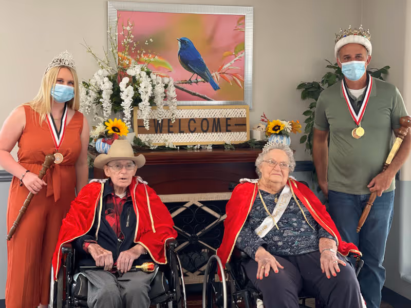 Two elderly individuals seated in wheelchairs wearing red capes and crowns, flanked by a younger woman and man standing on either side, both wearing crowns and holding wooden scepters. They are posing indoors in front of a fireplace decorated with flowers, a welcome sign, and a framed picture of a blue bird on a branch.