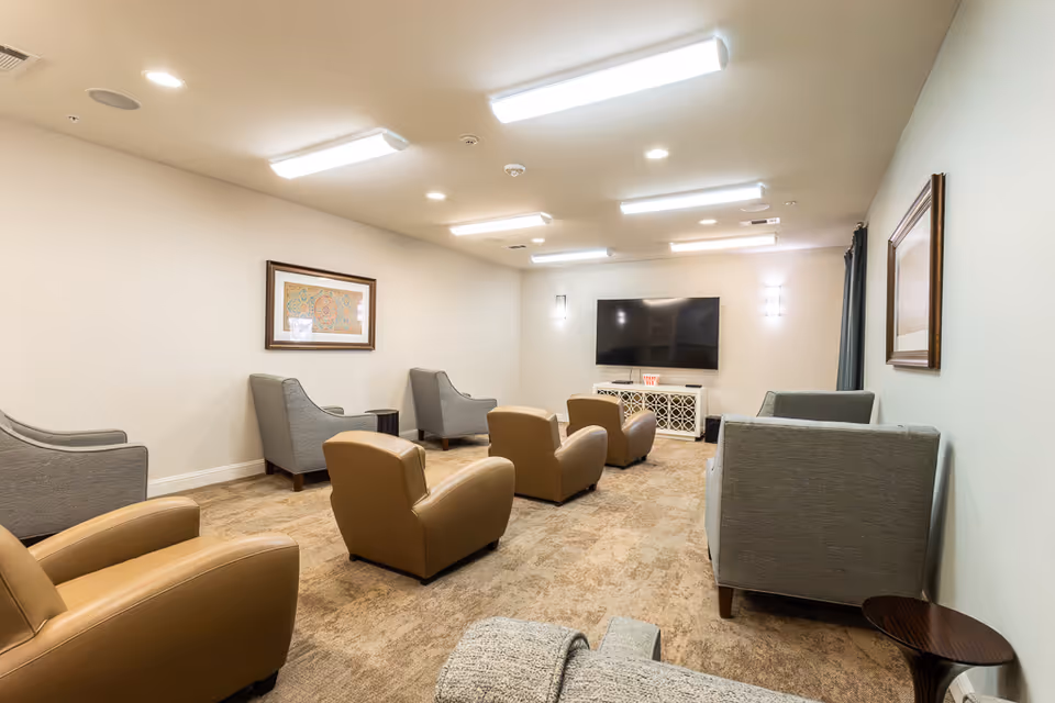 A cozy living room with several armchairs arranged facing a large flat-screen TV mounted on the wall. The room has beige carpet, neutral-colored walls, framed artwork, and bright ceiling lights.