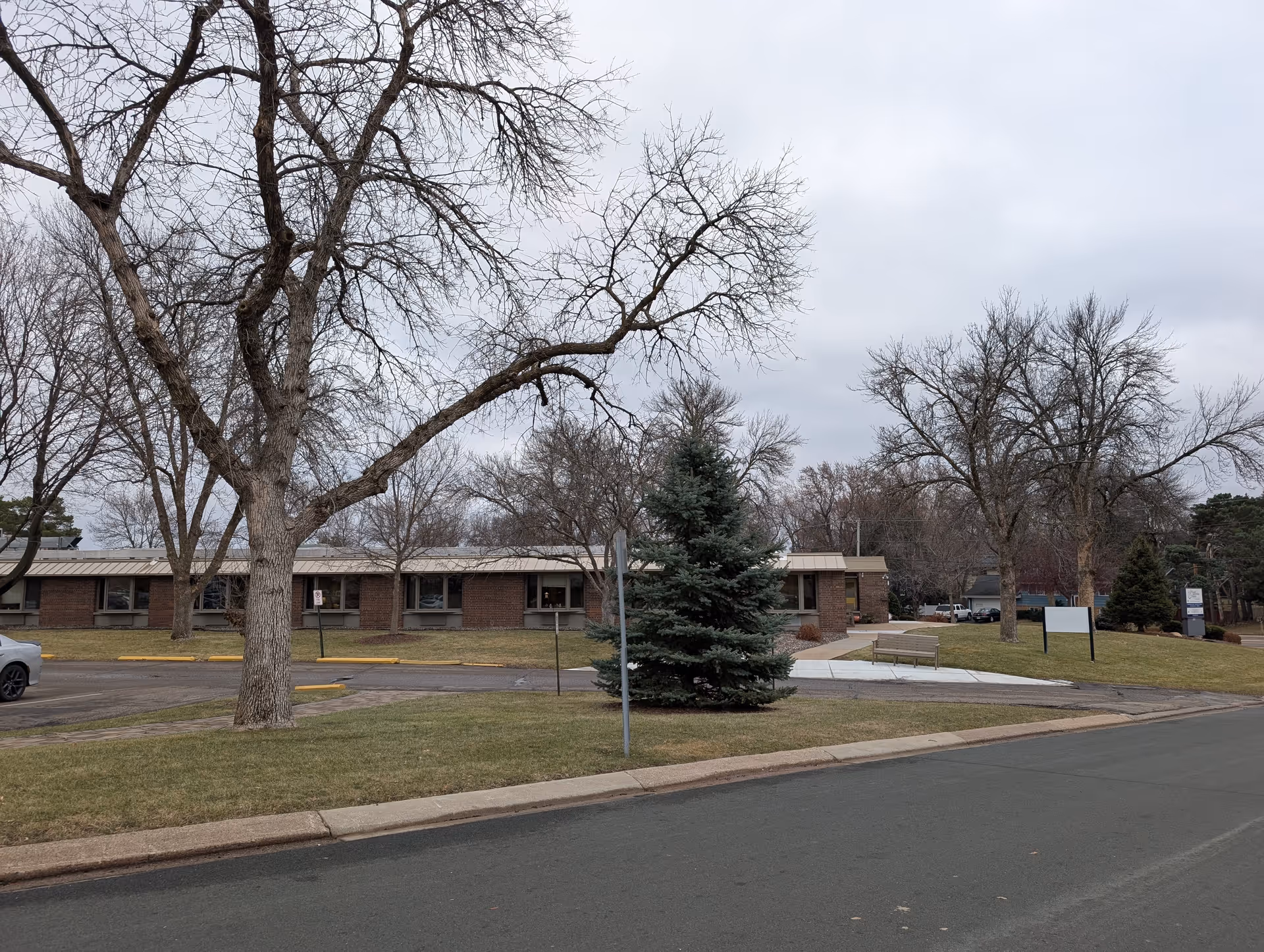 Low single-story brick senior living building seen from the street with leafless trees and an evergreen on a cloudy day.