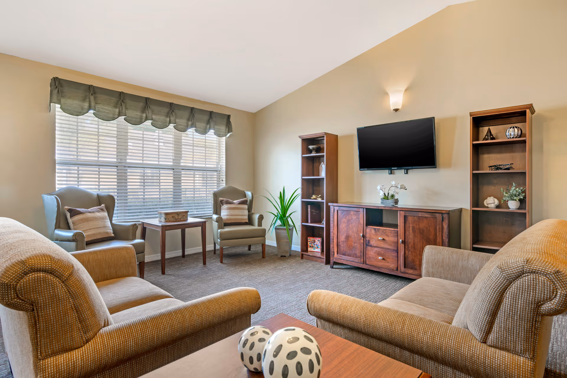 A cozy living room with two beige armchairs facing each other, two green wingback chairs with striped pillows near a large window with blinds and a valance, a wooden coffee table with decorative spheres, a wooden TV stand with a flat-screen TV mounted on the wall above it, and two wooden bookshelves with decorative items on either side of the TV stand. A potted plant is placed between the TV stand and one of the wingback chairs.