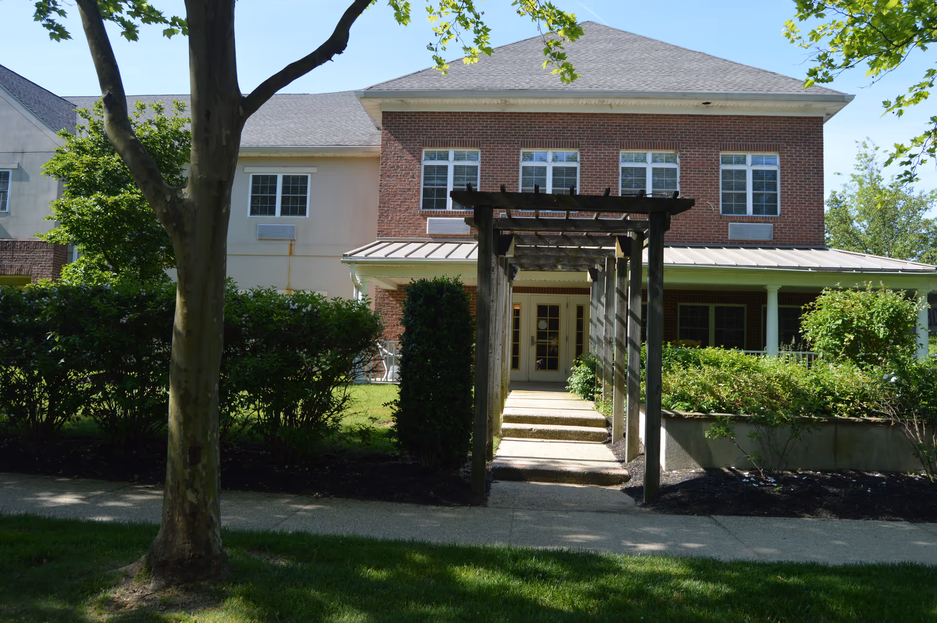 Exterior view of a two-story building with a brick facade and multiple windows. A wooden pergola leads to a door entrance surrounded by greenery and bushes. A tree and a sidewalk are visible in the foreground under a clear blue sky.