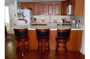 Interior view of a kitchen area with wooden cabinets, a white refrigerator covered with magnets and papers, a microwave, and a countertop with three wooden bar stools with black cushioned seats. The floor is wooden, and there is a doorway visible to the left.