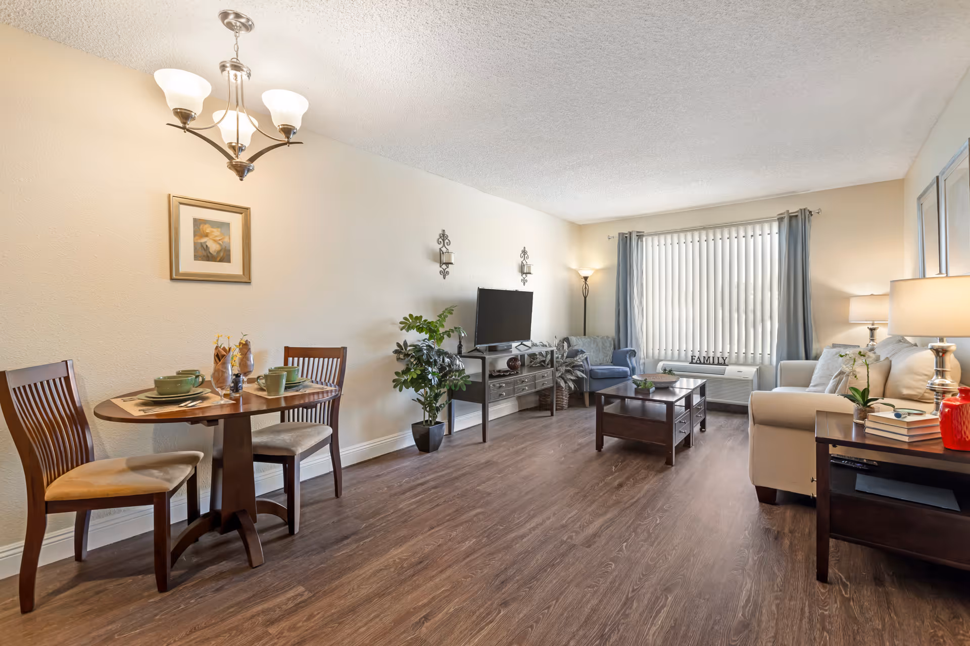 A cozy living room and dining area in a senior living facility. The room features a small round dining table set with green dishes and two wooden chairs on the left. The living area has a beige sofa, a blue armchair, a wooden coffee table, and a TV on a stand against the wall. There are decorative plants, wall sconces, a floor lamp, and a window with vertical blinds and blue curtains letting in natural light.