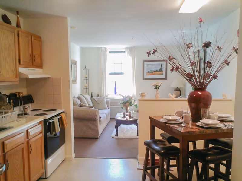 Open-plan apartment interior showing a small kitchen on the left, a dining table with place settings and a large vase, and a living room with a sofa and coffee table by a window.