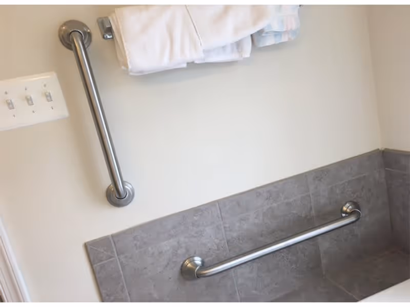 A bathroom wall with two stainless steel grab bars, one vertical and one horizontal, mounted above gray tiled walls. A towel rack with white towels is visible above the vertical grab bar, and a light switch panel with three switches is on the left side of the image.
