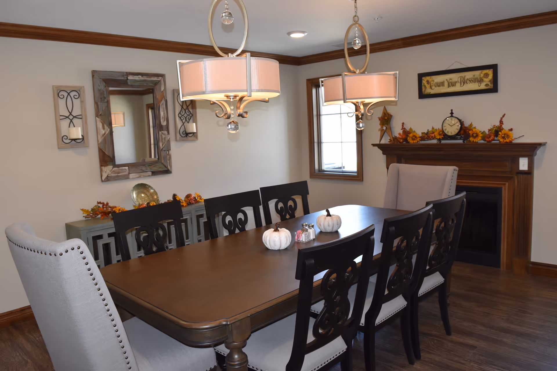 A dining room with a large wooden table surrounded by eight chairs, two of which have gray upholstery and nailhead trim. The table is decorated with two white pumpkins and a salt and pepper shaker set. Above the table hang two modern pendant lights with circular frames and crystal accents. The room features wood trim, a window, a fireplace mantel decorated with autumn-themed items and a clock, and a wall with a rustic mirror and candle sconces.