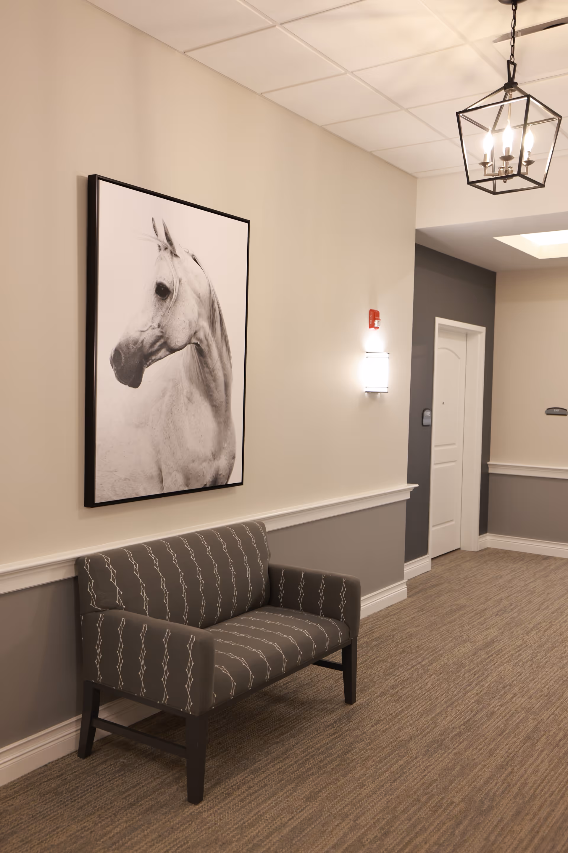 A hallway in a senior living facility with beige and gray walls, a patterned gray loveseat, a large black and white framed photograph of a horse on the wall, a ceiling light fixture, and a white door at the end of the hallway.