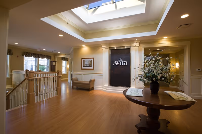 Interior view of a senior living facility lobby with wooden flooring, a round wooden table with a flower arrangement, a bench, framed artwork on the wall, a skylight, and a decorative wall feature with the word 'Avita'.