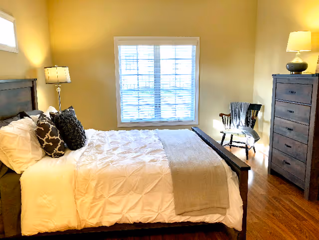 Sunlit bedroom with a neatly made bed, decorative pillows, a window with blinds, a wooden dresser and a chair.