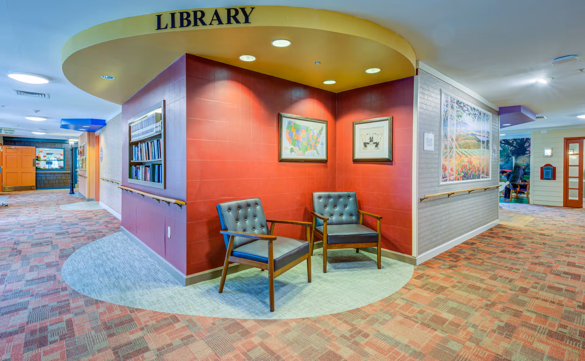 Interior view of a senior living facility hallway with a small library nook. The nook has a red wall with two framed pictures and two blue cushioned chairs. Above the nook, a yellow curved sign reads 'LIBRARY'. The hallway has patterned carpet and handrails along the walls, with additional artwork and doors visible further down the corridor.