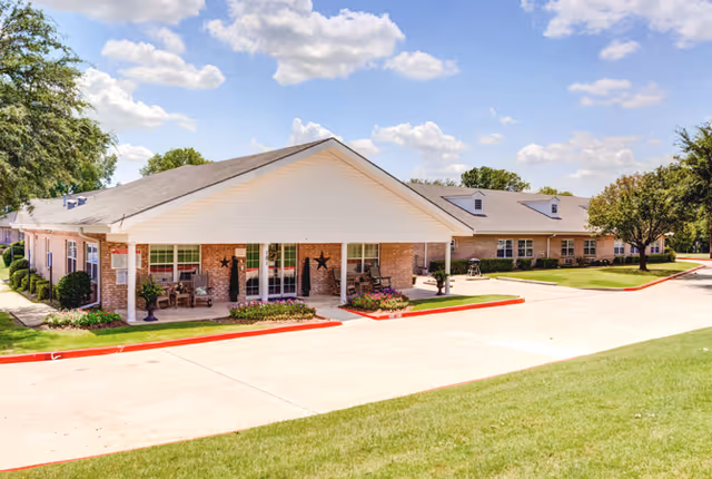 Exterior view of a single-story brick building with a white gabled roof, a covered porch with seating, and decorative stars on the walls. The building is surrounded by a well-maintained lawn, trees, and a paved driveway under a partly cloudy blue sky.