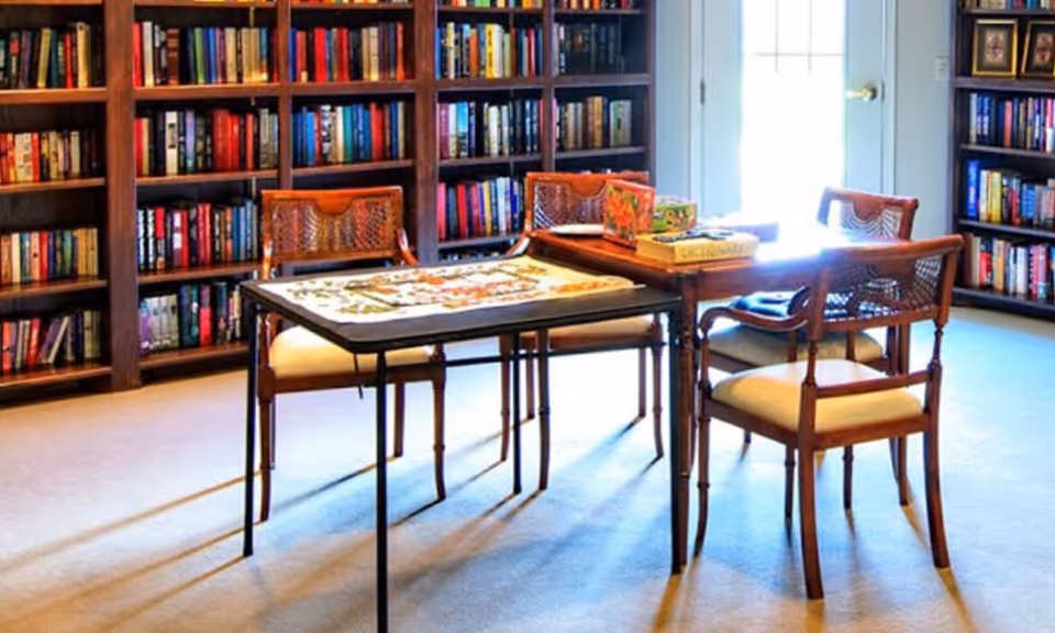 A bright common room with bookshelves lining the walls and a table with four chairs and a puzzle on top.
