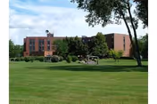 A large brick building with multiple windows surrounded by green trees and a well-maintained grassy lawn under a partly cloudy sky.