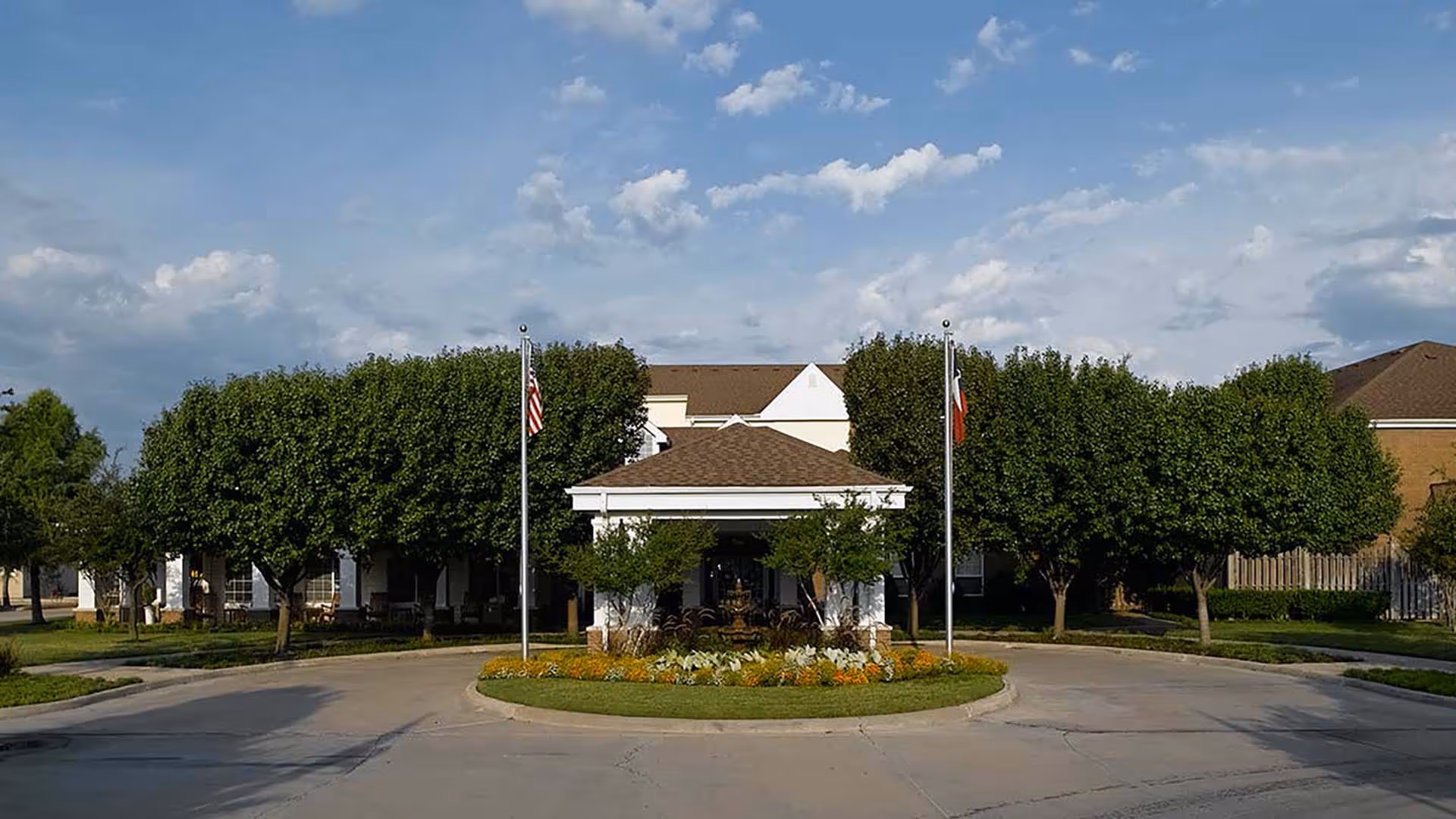 Front entrance of a senior living building with a circular driveway, flagpoles, neatly trimmed trees and landscaped planting under a blue sky.