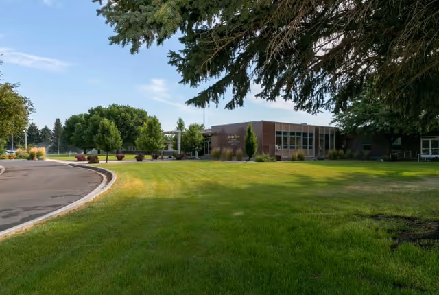Exterior view of Parke View Rehabilitation & Care Center showing a single-story brick building with large windows, surrounded by a well-maintained lawn, trees, and a curved driveway under a partly cloudy sky.