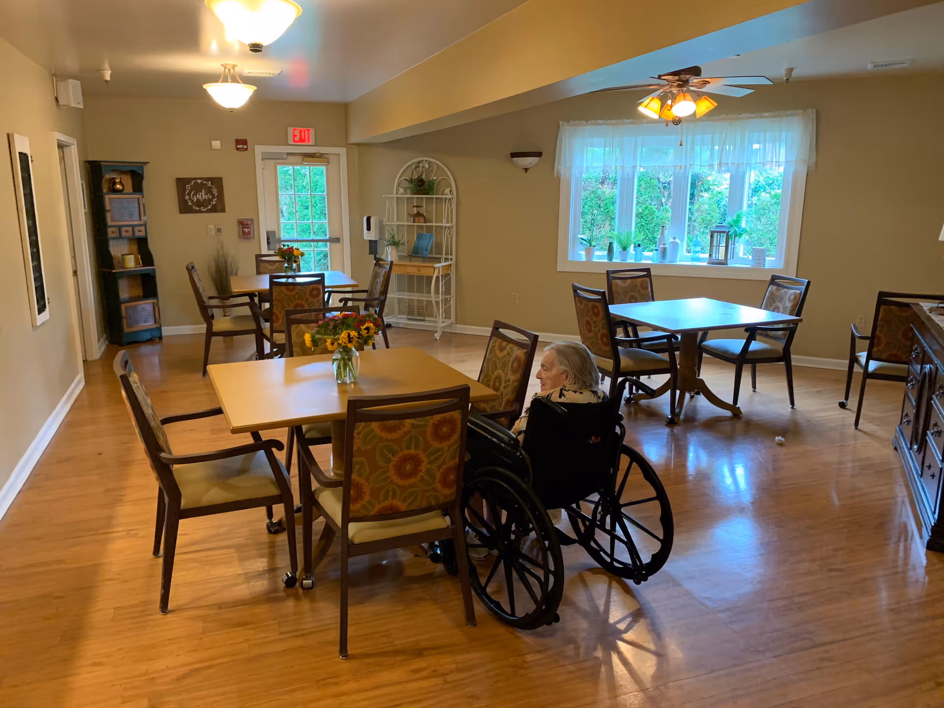 A dining room in a senior living facility with several wooden tables and chairs arranged neatly. A woman in a wheelchair is seated at one of the tables. The room has hardwood floors, beige walls, a large window with sheer curtains letting in natural light, and ceiling lights including a ceiling fan with lights. There are decorative items on a white shelf near the back door and a small cabinet on the right side.