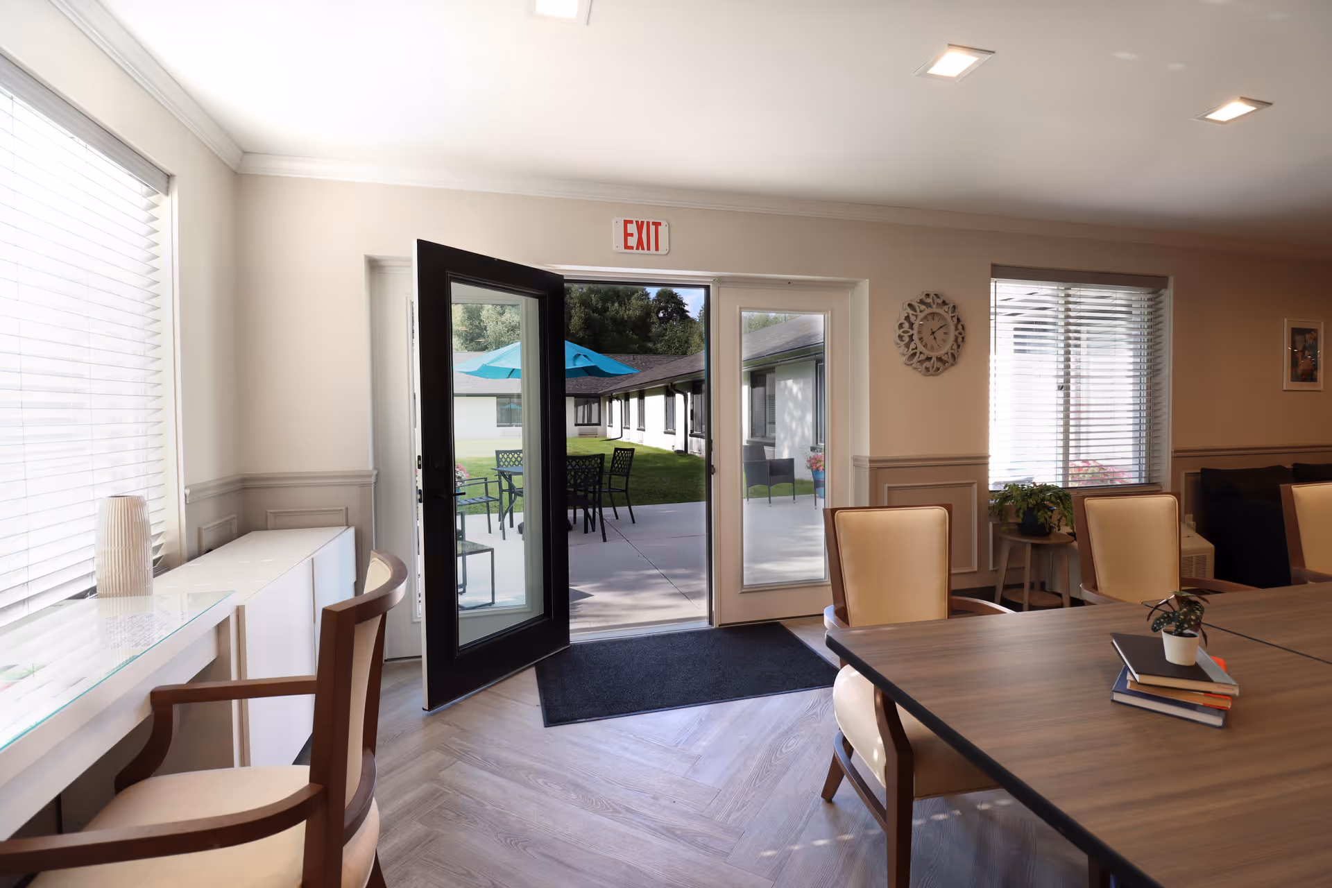 Communal dining area with tables and chairs and an open glass door leading to a patio with outdoor tables and umbrellas.