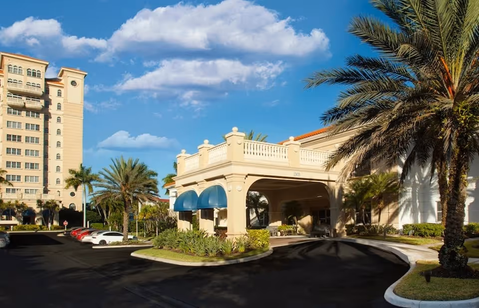 Exterior view of The Inn at Sarasota Bay Club showing a multi-story building with balconies, palm trees, a driveway with parked cars, and a covered entrance with blue awnings under a partly cloudy sky.