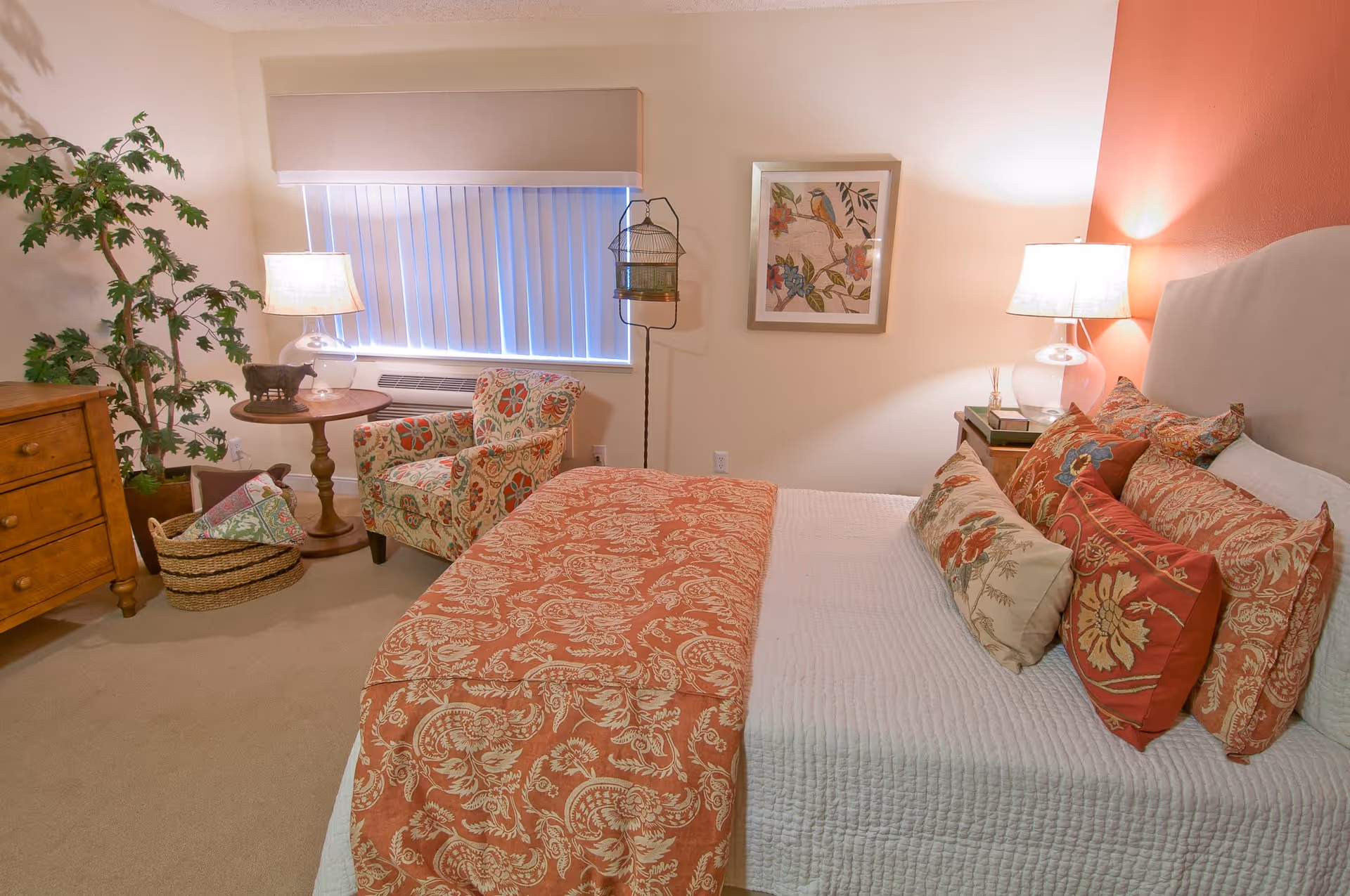 A cozy bedroom with a bed covered in a white quilt and multiple decorative pillows in red and beige patterns. There is a matching red and beige patterned throw blanket at the foot of the bed. To the right of the bed is a nightstand with a lamp. On the left side of the room, there is a wooden dresser, a round wooden side table with a lamp and a small decorative item, and a patterned armchair. A large potted plant is in the corner near the dresser. The window has vertical blinds and a beige valance. A framed picture of a bird and flowers hangs on the wall above the armchair.