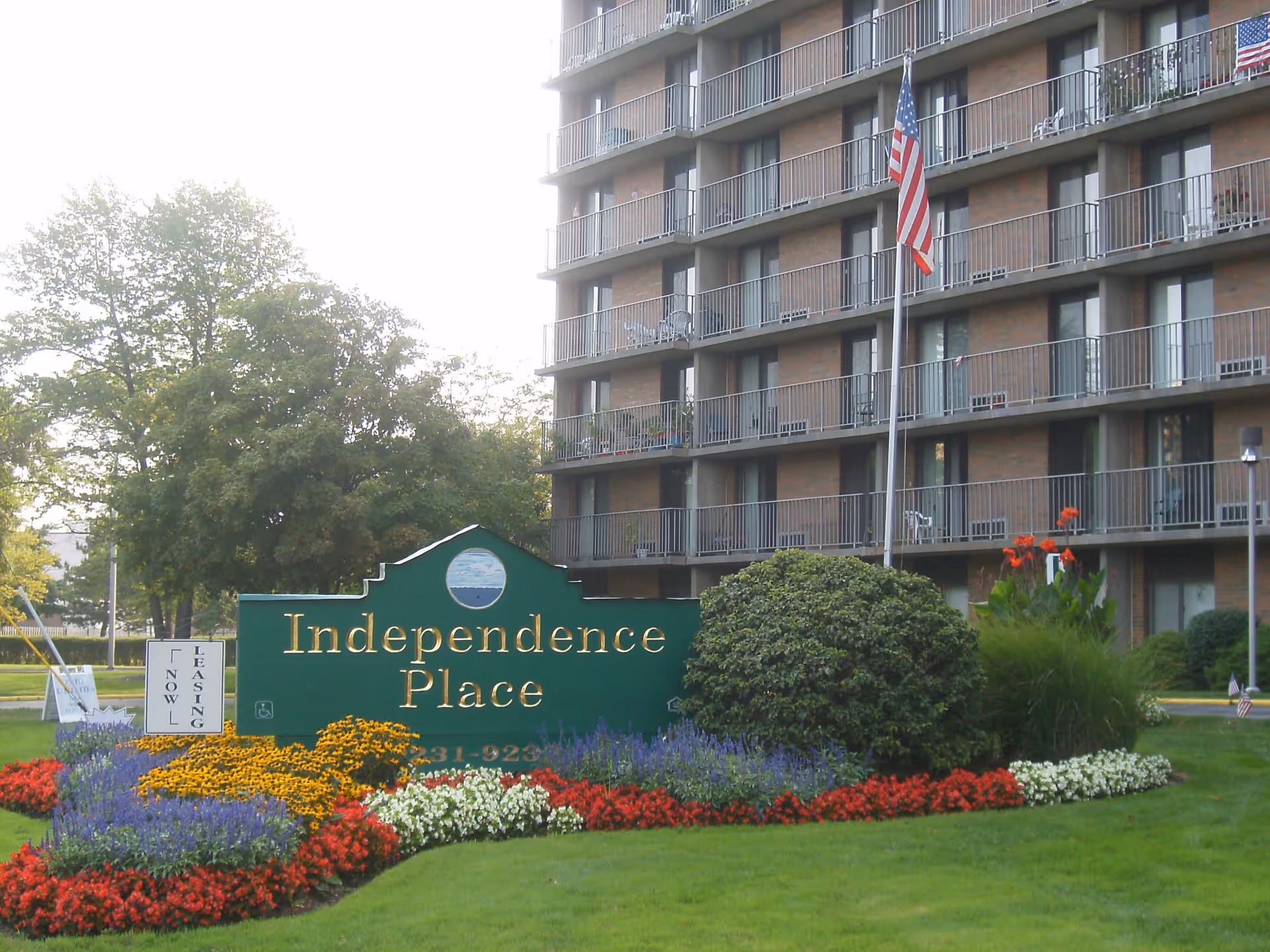 Outdoor view of Independence Place I facility showing a large green sign with gold lettering surrounded by colorful flower beds and greenery. Behind the sign is a multi-story building with balconies and an American flag on a flagpole.