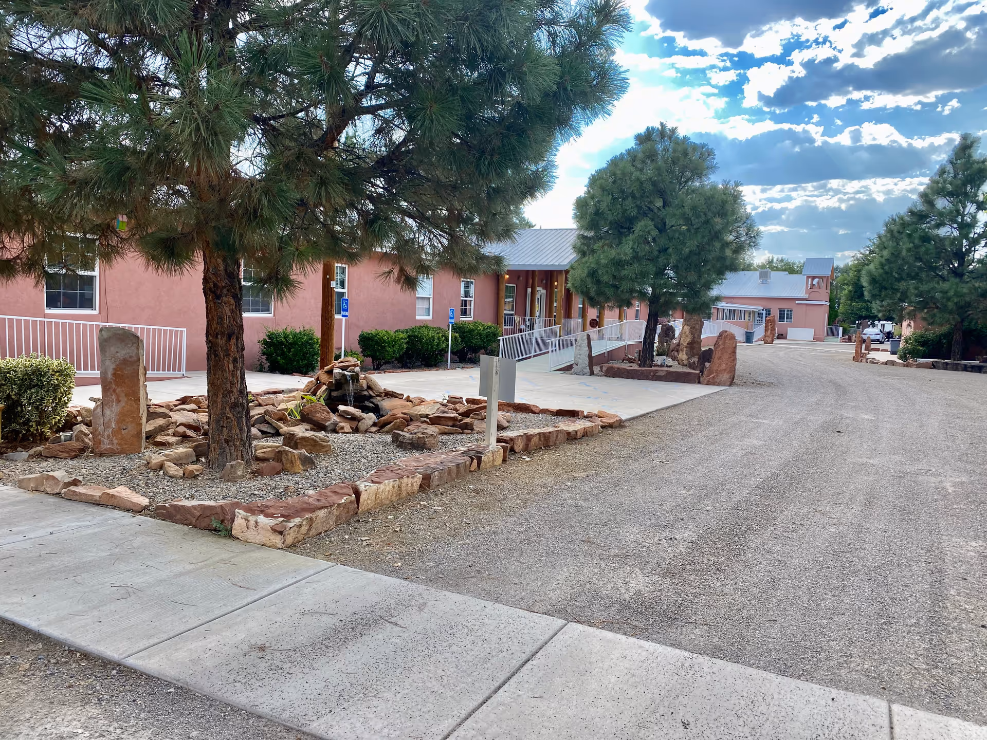 Outdoor view of Casa de Rosa Assisted Living facility showing a gravel driveway, landscaped area with rocks and trees, and a pink building with a metal roof in the background under a partly cloudy sky.