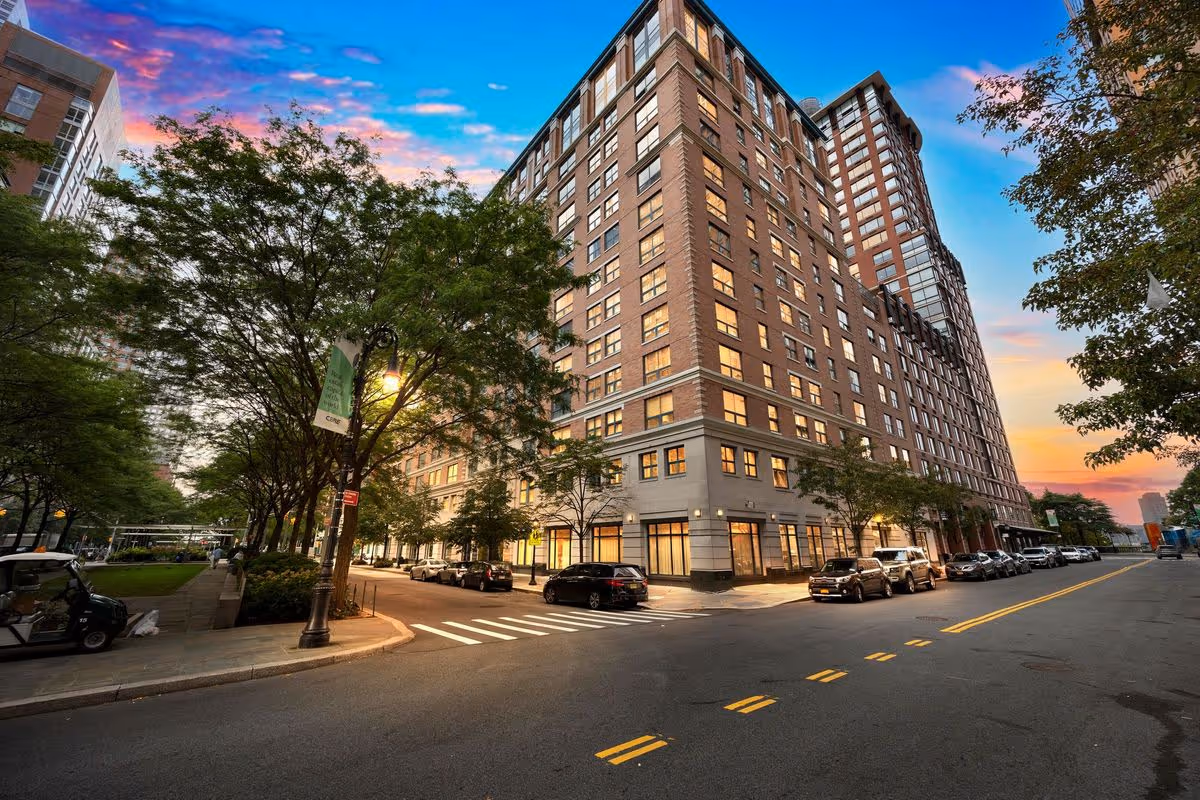 Street view of a large multi-story brick building at sunset with trees lining the sidewalk and cars parked along the street.