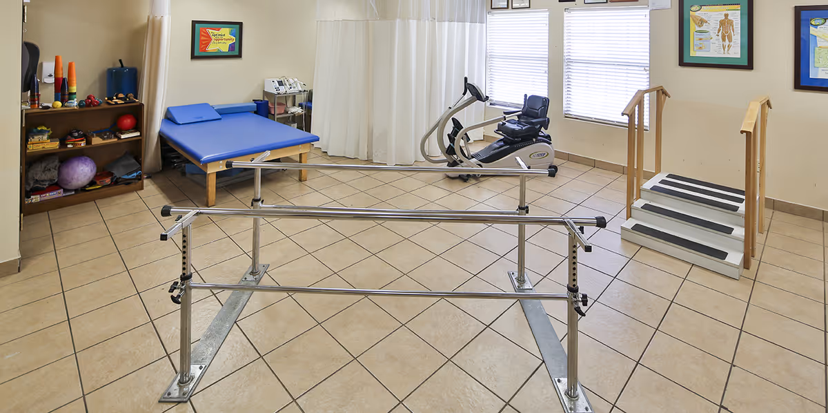 Physical therapy room with parallel bars for walking exercises, a blue padded therapy table, exercise equipment including a recumbent bike, a small set of stairs with handrails, and shelves with therapy tools and exercise balls. The room has tiled floors and windows with blinds.